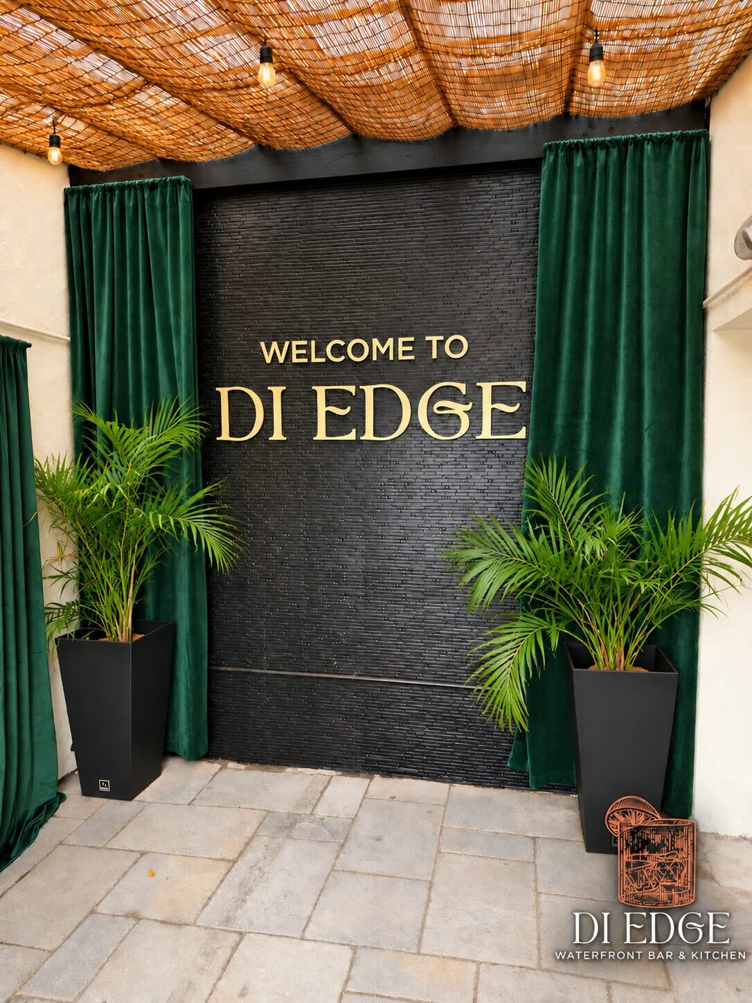 Gold lettering on black stone: Welcome to Di Edge, framed by emerald curtains and palms