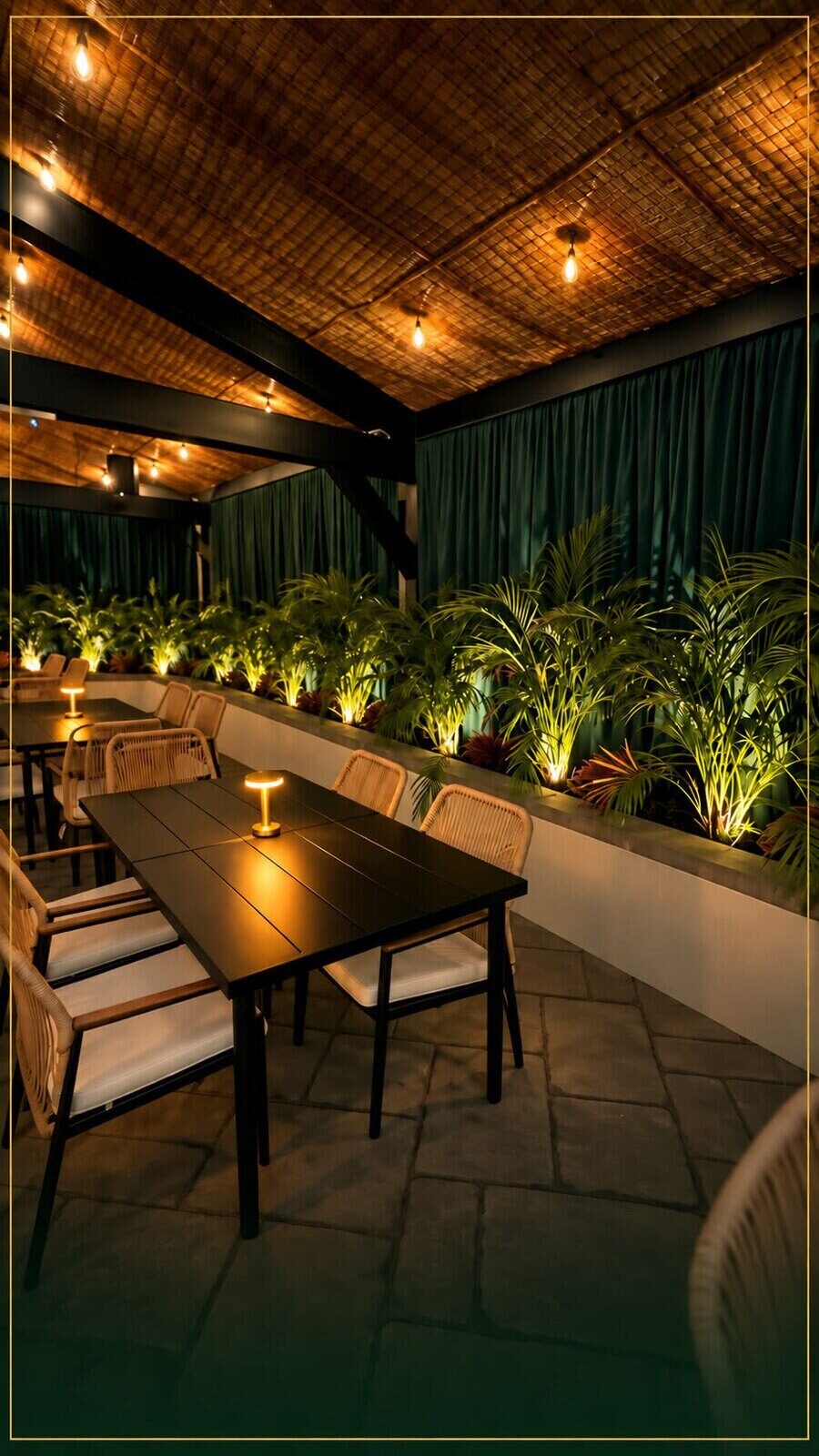 A private table with a brass lamp, palms lit from below, and emerald curtains